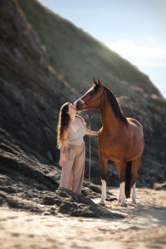 Cheval bai fait un bisou à sa propriétaire à la plage, entourés par des roches noires